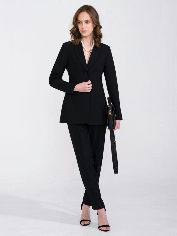 A stylish woman in a black suit poses elegantly in a studio setting, exuding confidence and fashion-forward vibes.